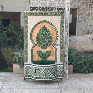 Puede incluir: Una fuente marroqu&iacute; decorativa con azulejos verdes y blancos. La fuente presenta un dise&ntilde;o de arco tradicional y un motivo floral central. La fuente est&aacute; sobre ruedas y tiene el texto "FARAH DECOR SOUADIA" en la parte inferior.