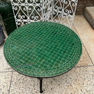 May include: A round green mosaic table with a black metal frame. The table top is made of small green tiles arranged in a geometric pattern. The table is sitting on a white tiled floor.