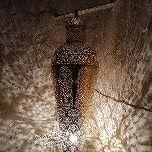 May include: A tall, gold-colored metal floor lamp with intricate cut-out designs. The lamp has a round base and a cylindrical shade with a pointed top. The lamp is lit and casting shadows on the wall behind it.Moroccan Brass Floor Lamp - Handmade Luxury Standing Light  - Oriental Boho Home Lighting Accent .