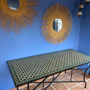 Op de afbeelding: Een rechthoekige tafel met een groen en beige betegeld blad en een zwart metalen frame. Twee ronde spiegels met stro-achtige franjes hangen aan een blauwe muur. De vloer is betegeld met terracotta vierkanten.