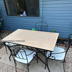 May include: A black wrought iron patio dining set with a square table and four chairs. The table top is made of beige mosaic tiles. The set is on a beige and brown patterned rug. The text "FARAH DECOR SOUDAGE" is visible on the table.