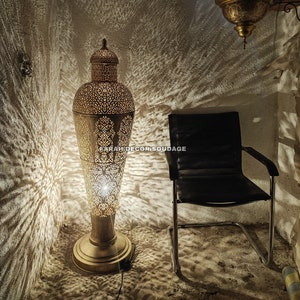 Op de afbeelding: Een hoge, sierlijke, goudkleurige metalen vloerlamp met ingewikkelde uitsnijdingen. De lamp is aan en werpt een warme gloed op de omgeving. Een zwarte leren stoel met een zilveren frame staat op de achtergrond.