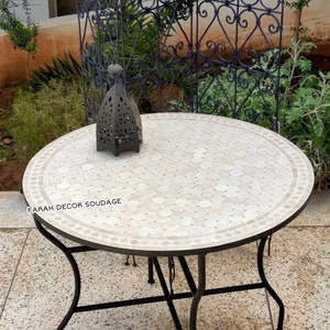Puede incluir: Una mesa redonda con una tapa de mosaico de azulejos blancos y un marco de metal negro. La mesa está sentada en un patio con una valla de metal decorativa en el fondo.
