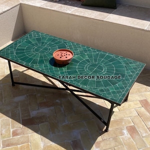 Op de afbeelding: Een groene mozaïek tegelblad salontafel met een zwart metalen frame. De tafel staat op een terras met een bakstenen vloer. De tafel heeft een kleine terracotta pot met een plant.