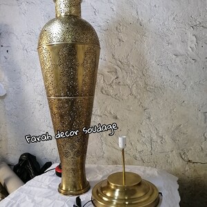K&ouml;nnte beinhalten: Eine hohe, goldfarbene Metalllampe mit filigranen Ausschnitten. Die Lampe hat einen runden Sockel und einen zylindrischen Schirm. Die Lampe steht auf einer wei&szlig;en Oberfl&auml;che. Der Text "Farah decor soudage" ist im Bild sichtbar.