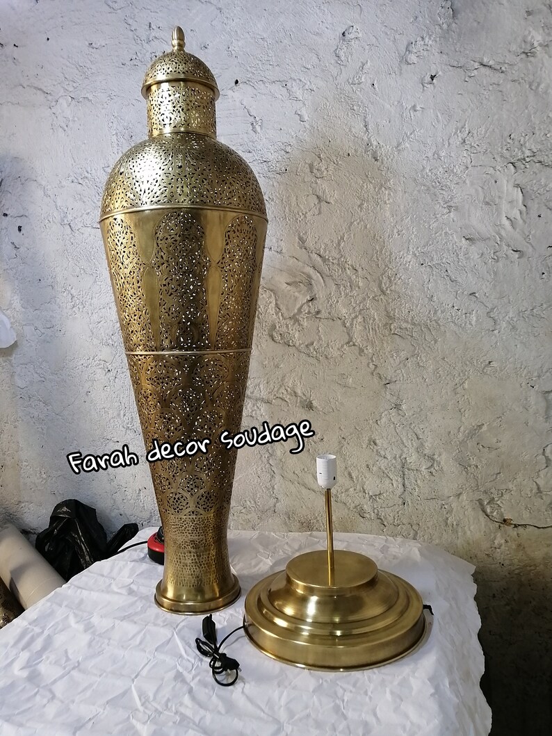 K&ouml;nnte beinhalten: Eine hohe, goldfarbene Metalllampe mit filigranen Ausschnitten. Die Lampe hat einen runden Sockel und einen zylindrischen Schirm. Die Lampe steht auf einer wei&szlig;en Oberfl&auml;che. Der Text "Farah decor soudage" ist im Bild sichtbar.