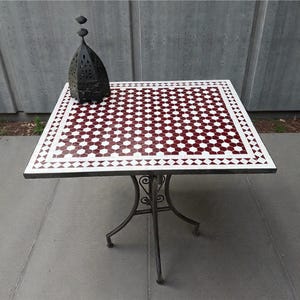 May include: Square outdoor table with a mosaic tile top in a red and white geometric pattern. The table has a wrought iron base with decorative scrollwork. A black metal lantern sits on the table.