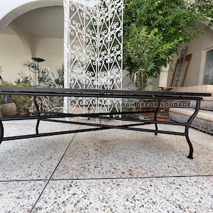 Puede incluir: Mesa de patio de metal negro con una tapa de vidrio rectangular. La mesa est&aacute; sentada en un patio de concreto con una puerta de metal blanco en el fondo.