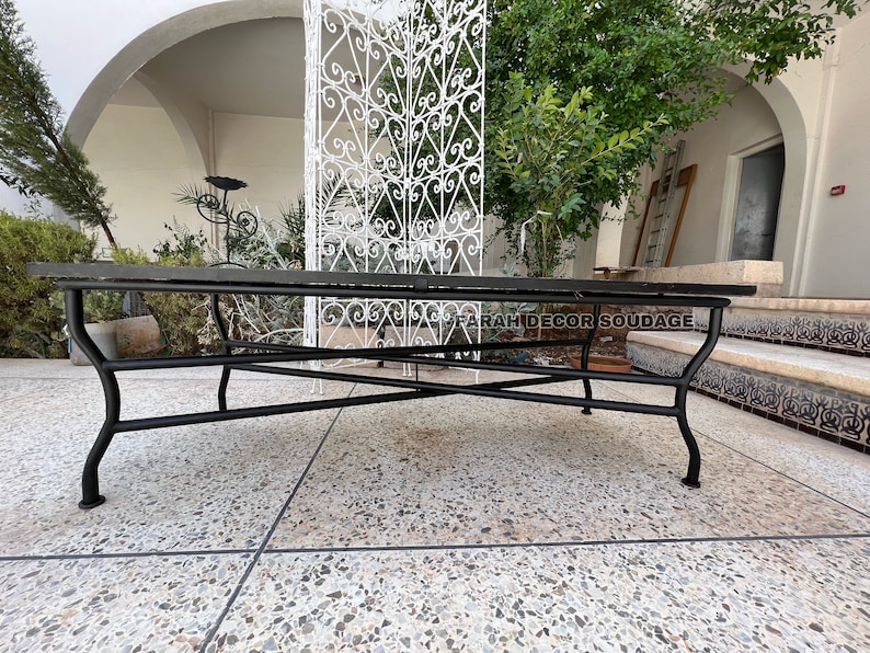 Puede incluir: Mesa de patio de metal negro con una tapa de vidrio rectangular. La mesa est&aacute; sentada en un patio de concreto con una puerta de metal blanco en el fondo.