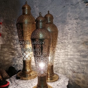 K&ouml;nnte beinhalten: Drei goldene Metall-Stehlampen mit filigranen Ausschnitten. Die Lampen sind beleuchtet und strahlen ein warmes Licht aus. Die Lampen stehen auf einer wei&szlig;en Oberfl&auml;che. "Farah decor soudaqe" ist auf der Oberfl&auml;che geschrieben.