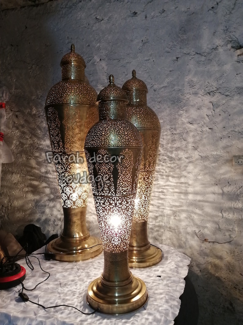 K&ouml;nnte beinhalten: Drei goldene Metall-Stehlampen mit filigranen Ausschnitten. Die Lampen sind beleuchtet und strahlen ein warmes Licht aus. Die Lampen stehen auf einer wei&szlig;en Oberfl&auml;che. "Farah decor soudaqe" ist auf der Oberfl&auml;che geschrieben.