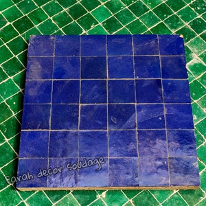 Può includere: Una lastra di piastrelle quadrata realizzata con piccole piastrelle di ceramica blu. Le piastrelle sono disposte a griglia. La lastra è appoggiata su una superficie piastrellata verde. Il testo "Farah decor soudadage" è scritto nella parte inferiore della lastra.