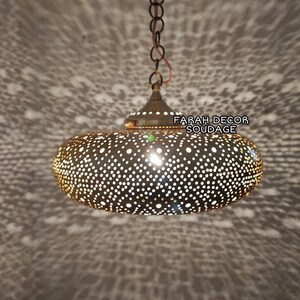 Może przedstawiać: Owalna, złota lampa wisząca z ozdobnym, skomplikowanym i szczeg&oacute;łowym wzorem małych otwor&oacute;w. Lampa jest zawieszona na łańcuchu i rzuca wzorzysty cień na sufit. Na lampie widoczny jest tekst "FARAH DECOR SOUDAGE".