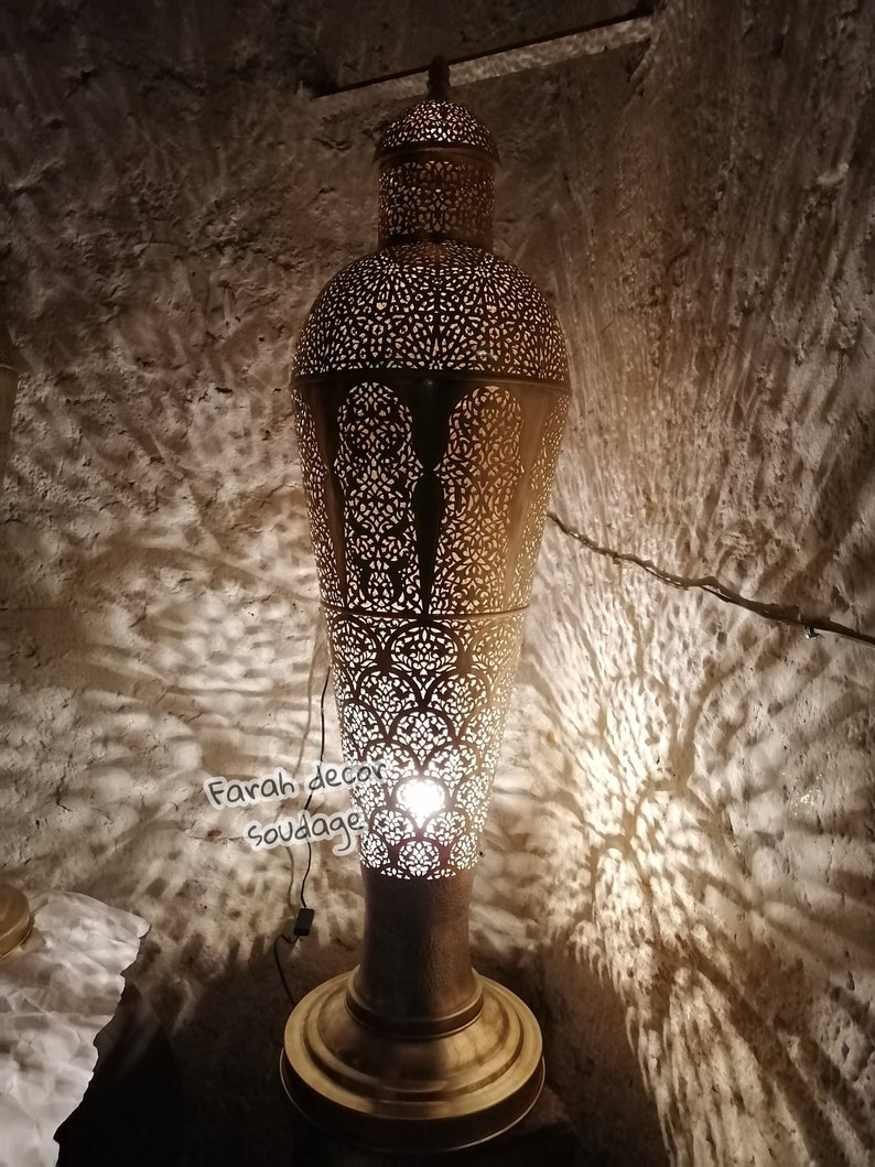 May include: A tall, gold-colored metal floor lamp with intricate cut-out designs. The lamp has a round base and a cylindrical shade with a pointed top. The lamp is lit and casting shadows on the wall behind it.Moroccan Brass Floor Lamp - Handmade Luxury Standing Light  - Oriental Boho Home Lighting Accent .