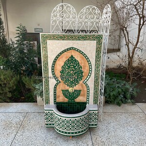 Puede incluir: Una fuente de pared decorativa con dise&ntilde;os de mosaico de azulejos verdes y blancos. La fuente presenta un arco tradicional marroqu&iacute; y una peque&ntilde;a cuenca en la parte inferior. El texto "FARAH DECOR SOUDAGE" es visible en la fuente.