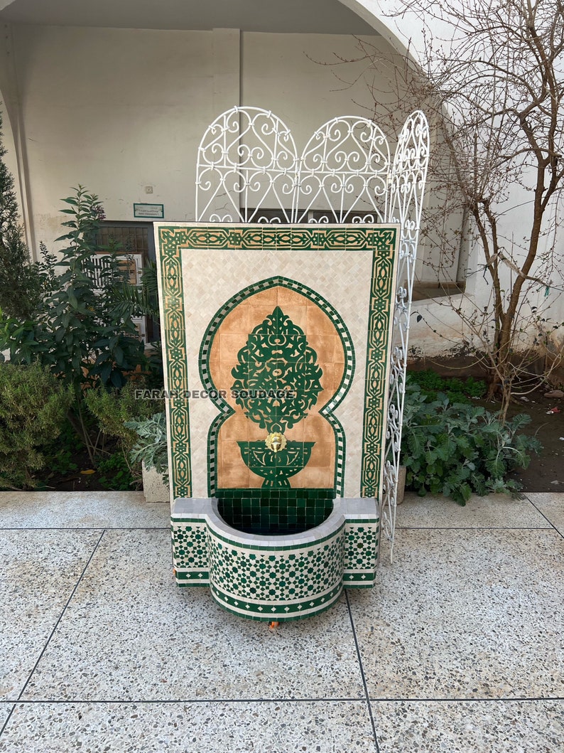 Puede incluir: Una fuente de pared decorativa con dise&ntilde;os de mosaico de azulejos verdes y blancos. La fuente presenta un arco tradicional marroqu&iacute; y una peque&ntilde;a cuenca en la parte inferior. El texto "FARAH DECOR SOUDAGE" es visible en la fuente.