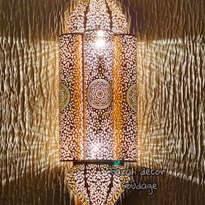 May include: A gold metal Moroccan lantern with intricate cutouts and a light shining through. The lantern is mounted on a wall.