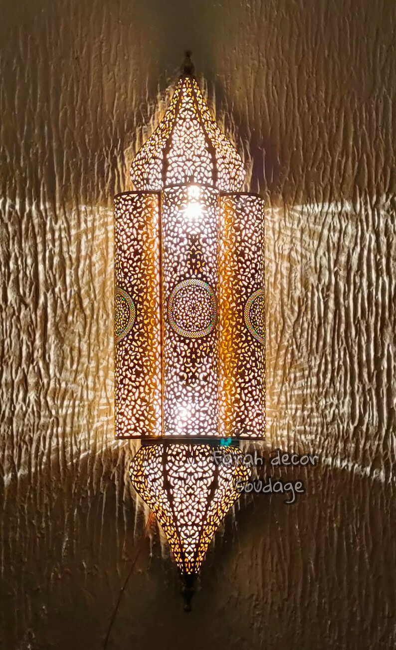 May include: A gold metal Moroccan lantern with intricate cutouts and a light shining through. The lantern is mounted on a wall.