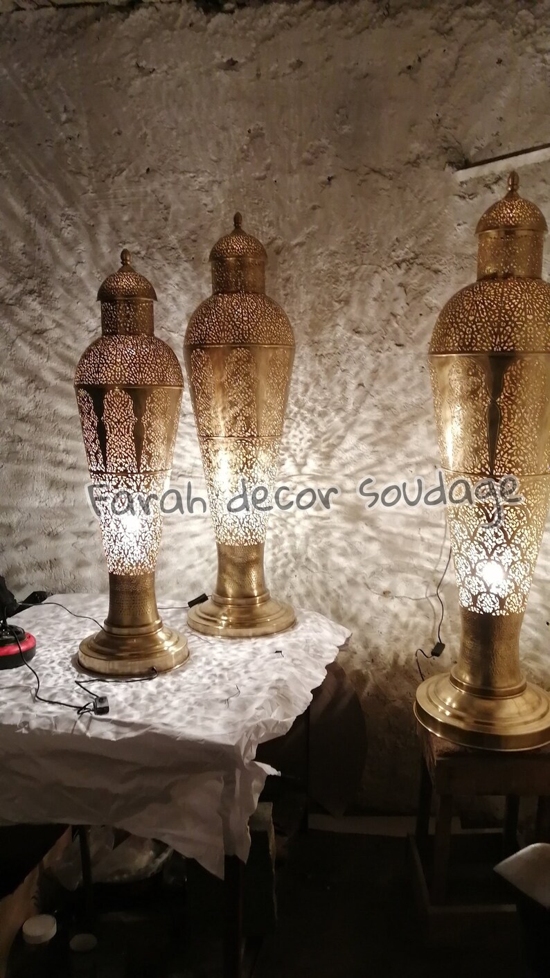 K&ouml;nnte beinhalten: Drei goldene Metall-Stehlampen mit filigranen Ausschnitten. Die Lampen sind beleuchtet und werfen Schatten auf die Wand hinter ihnen.