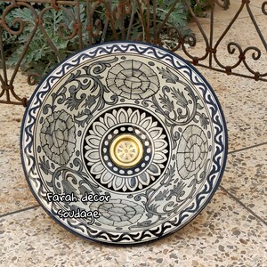 May include: A white ceramic bowl with a black and blue floral design. The bowl has a gold-coloured drain in the centre. The bowl is sitting on a tiled surface.