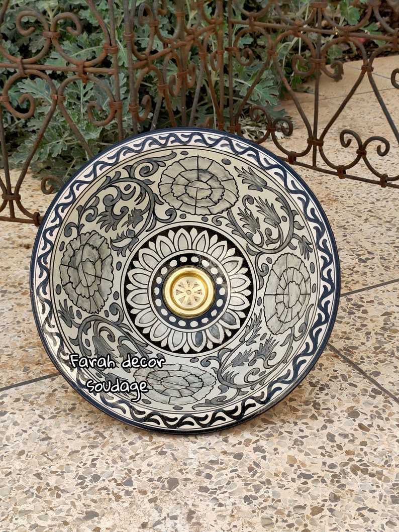 May include: A white ceramic bowl with a black and blue floral design. The bowl has a gold-coloured drain in the centre. The bowl is sitting on a tiled surface.