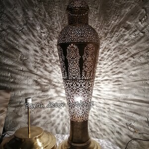 K&ouml;nnte beinhalten: Eine hohe, goldfarbene Metall-Stehlampe mit einem filigranen, ausgeschnittenen Design. Die Lampe hat einen runden Sockel und einen zylindrischen Schirm mit einer spitzen Oberseite. Die Lampe ist eingeschaltet und strahlt ein warmes Licht aus.