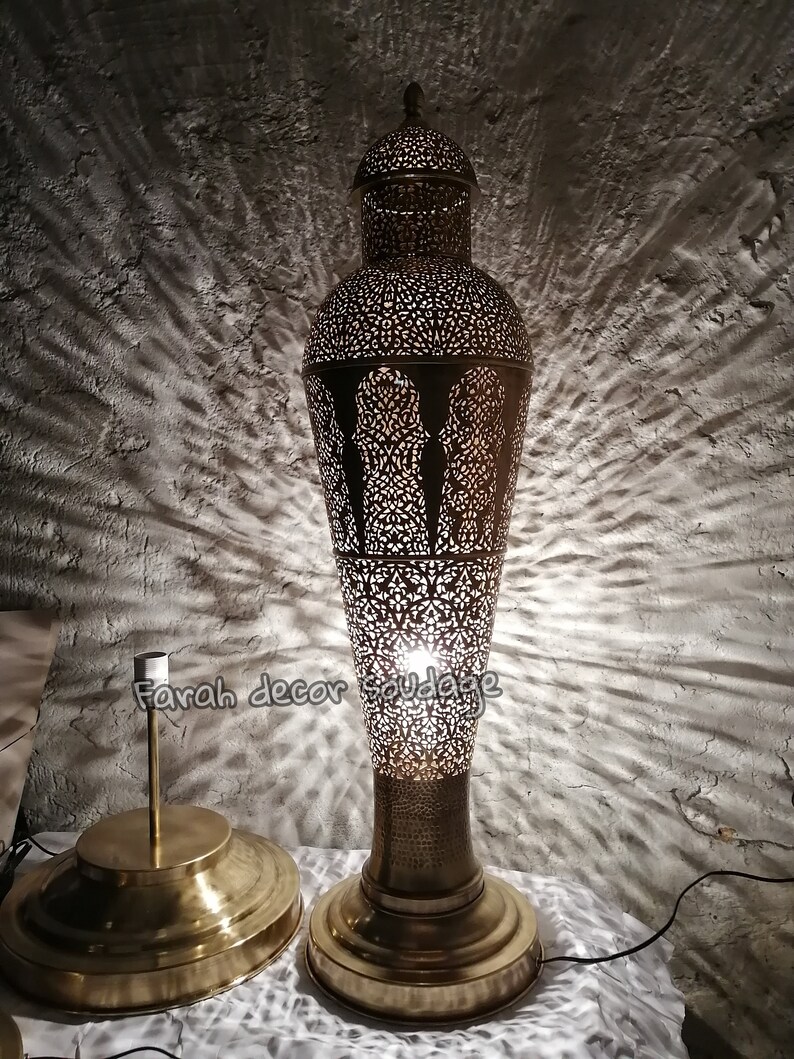 K&ouml;nnte beinhalten: Eine hohe, goldfarbene Metall-Stehlampe mit einem filigranen, ausgeschnittenen Design. Die Lampe hat einen runden Sockel und einen zylindrischen Schirm mit einer spitzen Oberseite. Die Lampe ist eingeschaltet und strahlt ein warmes Licht aus.