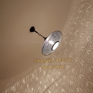 May include: A decorative, hanging pendant light with a perforated metal shade. The light fixture is suspended from the ceiling by a chain and emits a patterned light onto the surrounding surface. The text "FARAHD DECOR SOUDAGE" is visible.