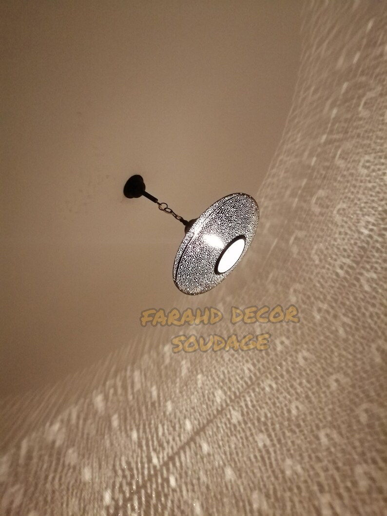 May include: A decorative, hanging pendant light with a perforated metal shade. The light fixture is suspended from the ceiling by a chain and emits a patterned light onto the surrounding surface. The text "FARAHD DECOR SOUDAGE" is visible.