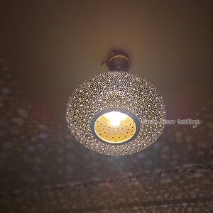May include: A decorative, round pendant light fixture with a perforated metal shade. The shade is dark with numerous small holes, allowing light to filter through. The fixture has a warm, inviting glow, casting patterned shadows on the ceiling.