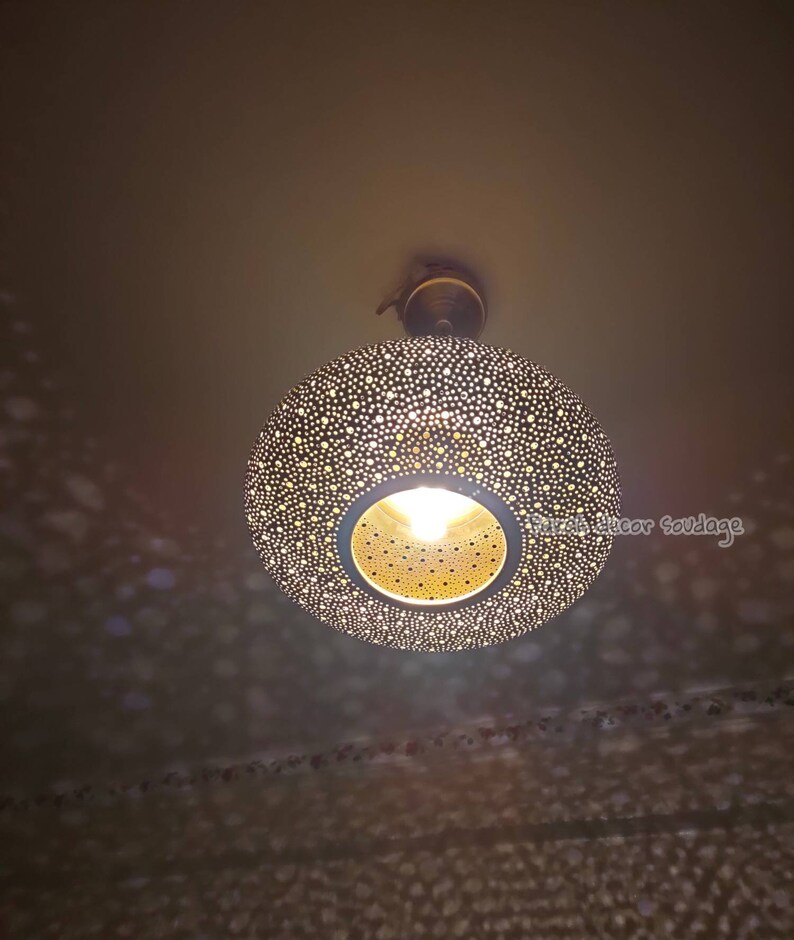 May include: A decorative, round pendant light fixture with a perforated metal shade. The shade is dark with numerous small holes, allowing light to filter through. The fixture has a warm, inviting glow, casting patterned shadows on the ceiling.