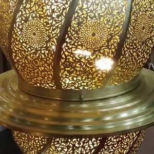 May include: A gold metal Moroccan lantern with intricate cut-out designs. The lantern is lit from within, casting a warm glow through the delicate patterns.