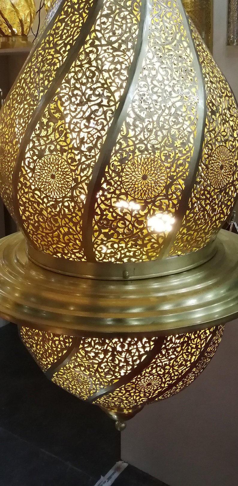 May include: A gold metal Moroccan lantern with intricate cut-out designs. The lantern is lit from within, casting a warm glow through the delicate patterns.