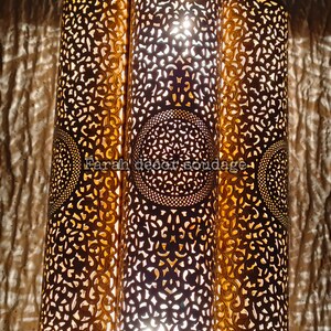 May include: A Moroccan-style pendant light fixture with three cylindrical shades. The shades are made of metal with intricate cut-out designs and a gold finish. The light is on and casting a warm glow.