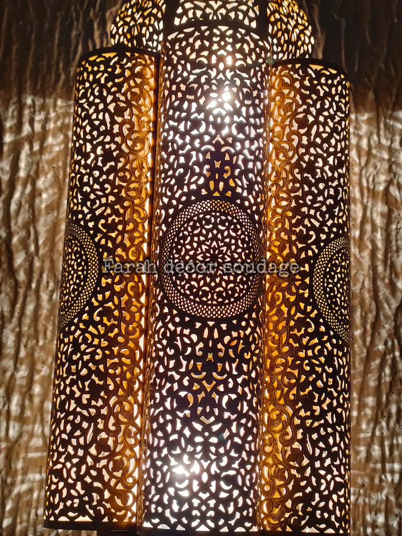 May include: A Moroccan-style pendant light fixture with three cylindrical shades. The shades are made of metal with intricate cut-out designs and a gold finish. The light is on and casting a warm glow.