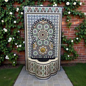 Op de afbeelding: Een decoratieve mozaïekfontein met ingewikkelde geometrische patronen in blauw, groen en goud. De fontein staat tegen een rode bakstenen muur met klimmende witte rozen. De fontein is ongeveer 2 meter hoog en 1 meter breed.