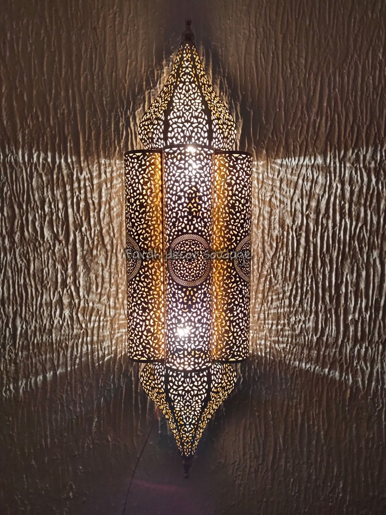 May include: A gold metal Moroccan lantern with intricate cut-out designs. The lantern is lit and casts a warm glow.Handmade Moroccan Wall Lamp | Brass Moroccan Sconce | Artisan Wall Lighting | Boho & Mediterranean Decor .