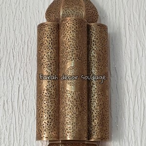 May include: A gold metal Moroccan style wall sconce with intricate cut-out designs. The sconce has three tiers and a pointed top.