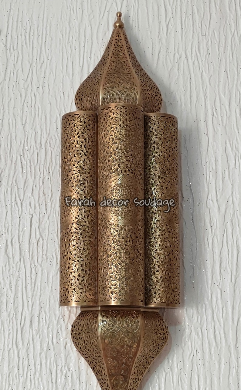 May include: A gold metal Moroccan style wall sconce with intricate cut-out designs. The sconce has three tiers and a pointed top.