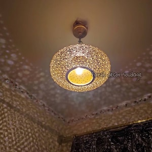 May include: A round, gold-colored pendant light fixture with a perforated design, casting patterned light onto the ceiling and walls. The light has a central bulb and is suspended from a ceiling mount.Round Oriental Moroccan Brass Lantern – Ceiling Pendant Light - Moorish lighting .