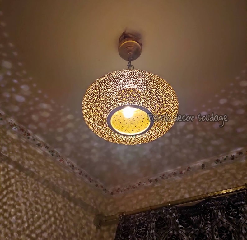 May include: A round, gold-colored pendant light fixture with a perforated design, casting patterned light onto the ceiling and walls. The light has a central bulb and is suspended from a ceiling mount.Round Oriental Moroccan Brass Lantern – Ceiling Pendant Light - Moorish lighting .