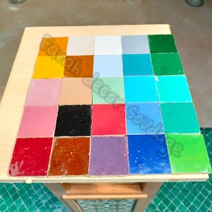 May include: A collection of 20 small square tiles in various colors, including yellow, red, blue, green, and purple. The tiles are arranged in a grid pattern on a wooden surface.