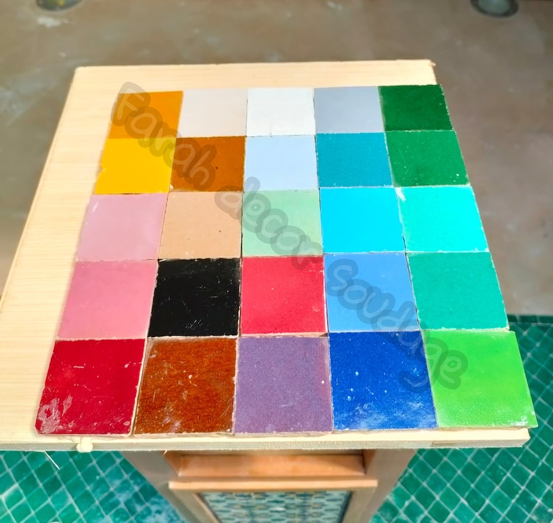 May include: A collection of 20 small square tiles in various colors, including yellow, red, blue, green, and purple. The tiles are arranged in a grid pattern on a wooden surface.