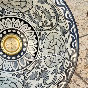 May include: A white ceramic bowl with a black and grey floral design. The bowl has a gold rim and a black and white border. The text "Farah decor soudaage" is written on the bowl.