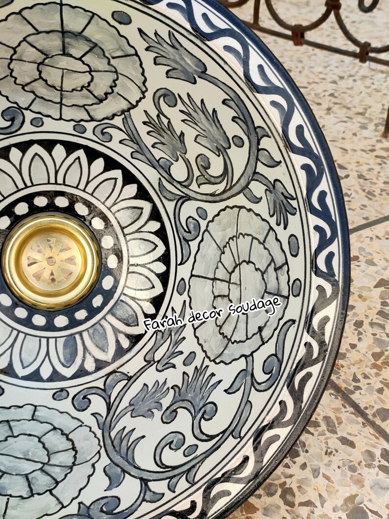 May include: A white ceramic bowl with a black and grey floral design. The bowl has a gold rim and a black and white border. The text "Farah decor soudaage" is written on the bowl.