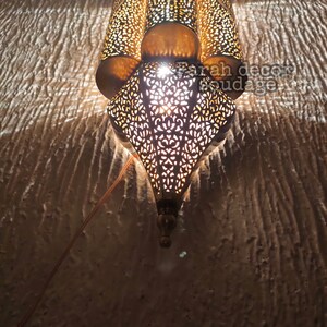May include: A gold metal Moroccan style lantern with intricate cut-out designs. The lantern is lit and casting a warm glow.