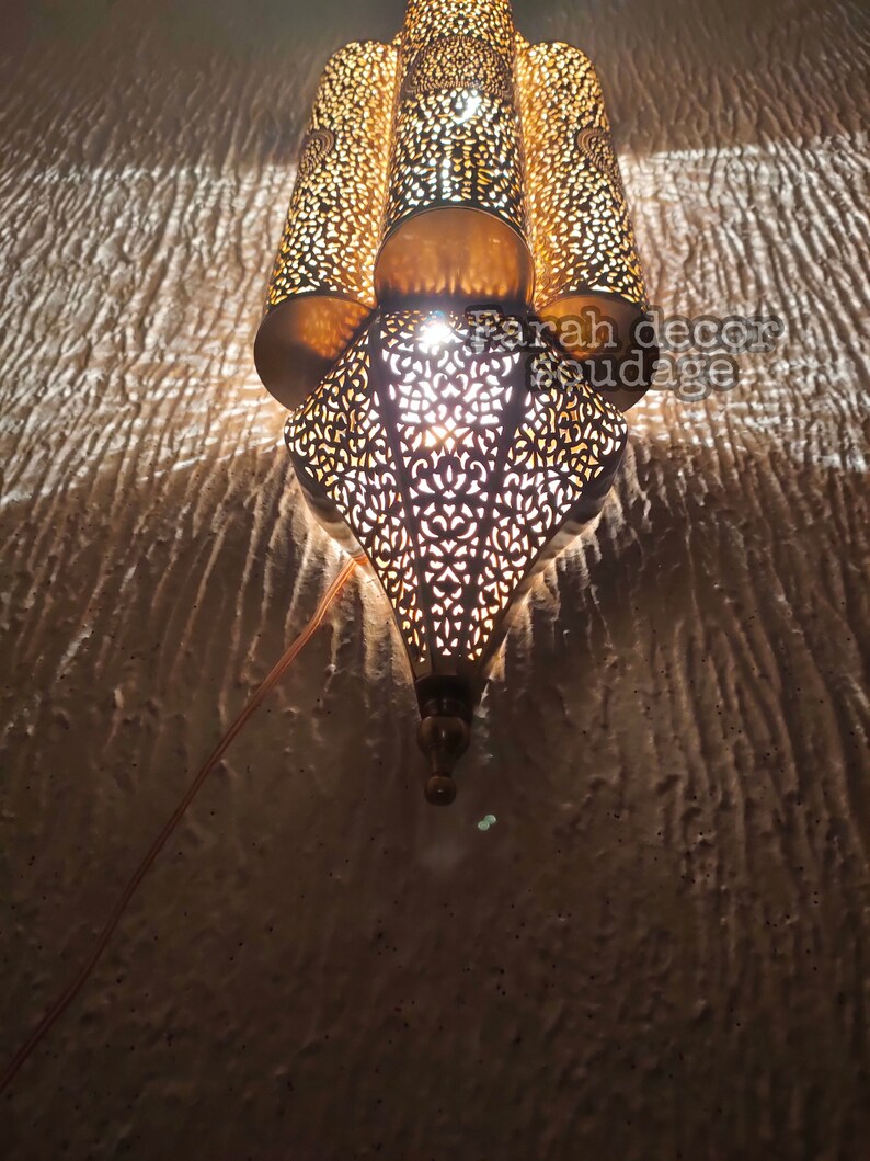 May include: A gold metal Moroccan style lantern with intricate cut-out designs. The lantern is lit and casting a warm glow.