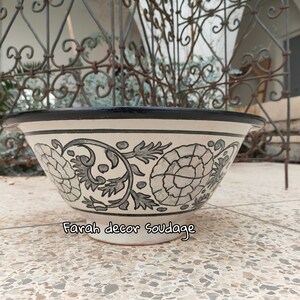 May include: A white ceramic bowl with a black rim and a floral design in grey and black. The bowl is sitting on a white tiled surface. The text "Farah decor soudaage" is visible on the bottom of the bowl.