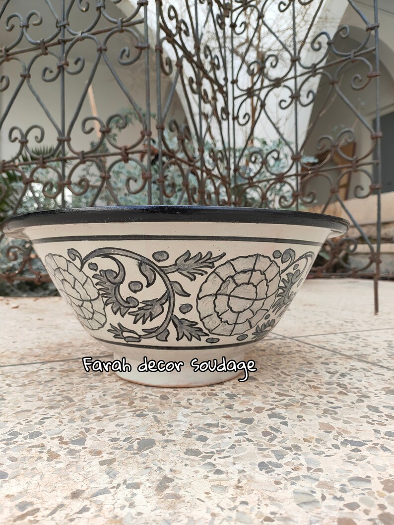 May include: A white ceramic bowl with a black rim and a floral design in grey and black. The bowl is sitting on a white tiled surface. The text "Farah decor soudaage" is visible on the bottom of the bowl.