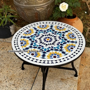 Pode incluir: Uma mesa redonda de mosaico com fundo branco e um padrão geométrico azul, amarelo e vermelho. A mesa tem uma estrutura de metal preto com três pernas.